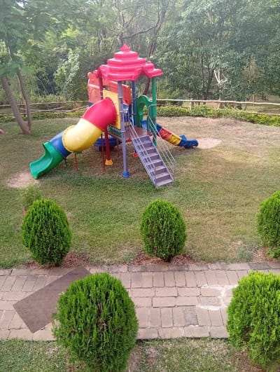 A playground structure, featuring slides and stairs, stands in a grassy area. Green bushes line a brick pathway. Trees form a backdrop behind a wooden fence. The slide is multi-colored with red, yellow, and green sections. The playground itself is painted in bright primary colors.