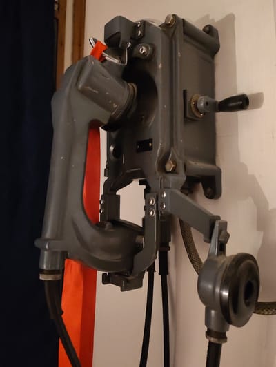 A gray, vintage, military-style telephone is mounted on a wall. The phone features a hand crank on the right side, a handset resting in a cradle, and a separate earpiece hanging below. Wires and cables connect the various components. An orange ribbon or strap is visible behind the handset. The location cannot be determined from the image.