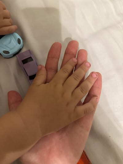 A close-up overhead shot shows two hands, one small and one large, placed together on a white, textured surface, possibly a bedsheet. The smaller hand is on top, with its fingers extended across the larger hand's palm. The child's fingernails appear dirty. Two toy cars, a light blue one and a lavender one, are visible on the surface to the left of the hands. The image provides no information about the city of Malinovka, Russia.