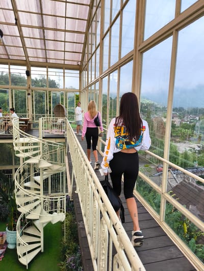 The image shows the interior of a glass-enclosed structure in Candikuning, Indonesia. Two women are walking away from the camera on a wooden bridge with a white railing, toward two other people. One woman wears a white long-sleeved shirt with a colorful graphic on the back and black leggings, carrying a black backpack. The other woman wears a pink hooded jacket and black leggings. To the left, a white spiral staircase descends to a lower level with plants. Other people are seated at a table in the background. Outside the glass walls, houses and greenery are visible.