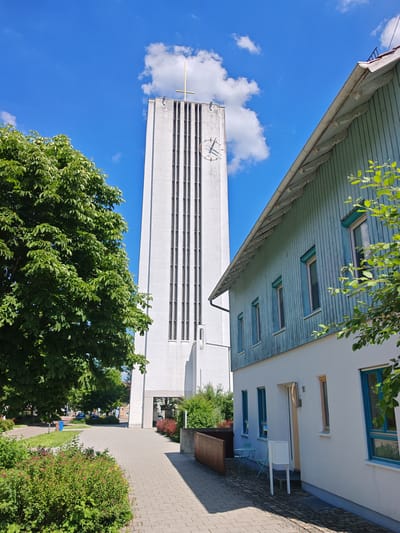 The image depicts a modern white bell tower, topped with a golden cross, adjacent to a pale blue wooden-sided building. The bell tower features vertical slits and a circular clock face. In the foreground, a paved walkway is bordered by green bushes and trees. The scene appears to be located in Memmingen, Germany, with a residential or community building next to the church tower. The sky is bright blue with scattered white clouds.