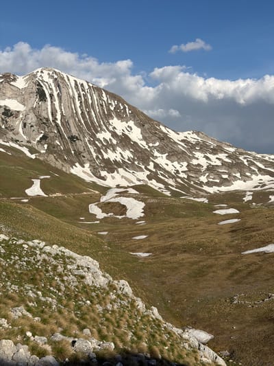 The image shows a mountain landscape in Boričje, Montenegro. The left side of the image features a steep, rocky mountain face with alternating layers of rock and snow. The right side of the image shows a grassy slope with scattered rocks and patches of snow. The sky is visible in the upper left corner and contains both blue sky and white and grey clouds.