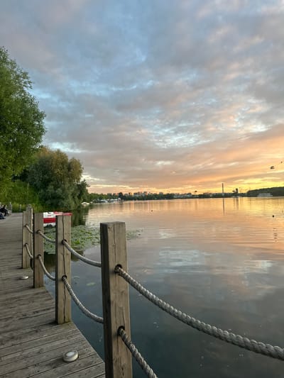 The image shows a wooden pier on a body of water, likely a lake or river, with a city skyline in the distance. The time of day appears to be sunset or sunrise, as indicated by the warm hues in the sky and the reflections on the water. The weather is cloudy, with a mix of soft, diffused light and darker clouds.
On the pier, there are thick ropes used as railings, strung through wooden posts. Part of a red and white boat is visible tied to the pier. The water is calm, with lily pads floating on the surface. Trees and green vegetation line the shore, and the distant city has several buildings, including a prominent chimney. A few people are seated on the pier in the foreground, seemingly enjoying the view. The overall mood is peaceful and serene, with a sense of calm at the end of the day. There is no visible text in the image.