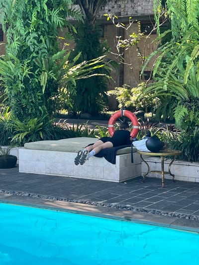 The image depicts a person sitting on a tiled bench next to a swimming pool. The pool's water is bright blue. Dense green vegetation surrounds the area. A life preserver is propped against the back of the bench. A small table with a black helmet on it sits nearby. The scene is located in Petulu, Indonesia.