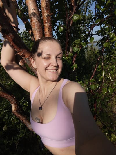 A young woman with brown hair pulled back poses for a selfie, leaning against a tree with reddish-brown bark. She is wearing a light pink tank top and a silver necklace with a dark pendant. A tattoo is visible on her chest. Sunlight dapples her face and arm, and she is smiling with her eyes partially closed. The background consists of lush green foliage and a bright blue sky, suggesting a warm, sunny day. In the distance, a body of water can be seen. The woman appears relaxed and happy, enjoying the outdoors. The overall scene is one of natural beauty and personal contentment. There is no visible text in the image.