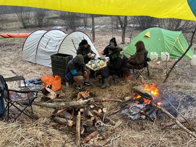 The image depicts a campsite scene, likely in a natural environment near Zabor'e, Russia. The foreground shows a campfire with burning logs and a pile of additional firewood. A grill sits over some coals, possibly for cooking food. In the center, five people are gathered around a small table, which is covered with food, drinks, and various containers. They appear to be eating or preparing food. To the left, there is a collapsible camping chair and a crate with bags. In the background, two tents are visible: a larger white tent and a smaller green tent. Beyond the tents, there is a sparse wooded area with signs of snow on the ground. An orange inflatable boat also lies to the left. A yellow and blue tarp is suspended above the people and the table.