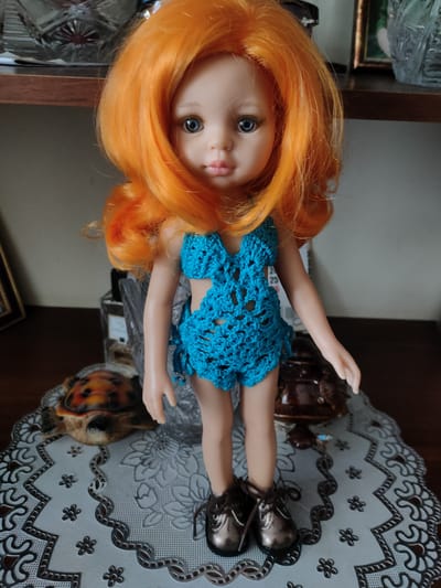 This media file features a medium shot of a doll with bright orange hair, blue eyes, and fair skin. The doll is wearing a crocheted turquoise swimsuit and metallic bronze lace-up shoes. It stands on a white crocheted doily placed on a wooden surface.
In the background, to the left of the doll, is a decorative glass object on a wooden shelf, and to the right, another decorative object. Further in the background, on the shelf, are framed pictures and more decorative glass items. Behind the doll, on the wooden surface, is a small decorative turtle figurine. The lighting suggests an indoor setting, possibly during the day, with artificial light contributing to the overall illumination. There is no visible text in the media file. The overall scene depicts a still life arrangement with a doll as the central subject. The location context is Salavat, Russia.