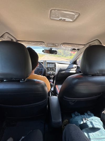 The image depicts the interior of a vehicle, viewed from a rear passenger seat looking forward, while in motion in Panglao, Philippines. The driver, a person with dark hair wearing a yellow-orange t-shirt, is visible from behind at the steering wheel. A mobile phone displaying a map is mounted to the right of the steering wheel. The car's dashboard features a central infotainment screen showing "92.7" and climate controls. A small black device, possibly a dashcam, is mounted near the rearview mirror. The sun visors remain wrapped in clear plastic. Black leather-like seats are visible, including the front passenger headrest and the back of the driver's seat with red trim. In the rear passenger footwell on the right, a teal/light blue backpack rests on the floor next to a foot wearing a brown Croc-style shoe. The exterior environment, visible through the windshield, shows a paved road lined with dense green vegetation under a bright sky, suggesting daytime. Power lines are visible in the distance on the left.