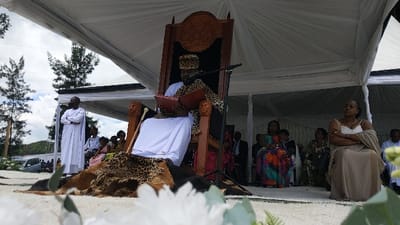 A man sits in a large, ornate wooden chair, reading a book. He wears a white robe, a leopard skin vest, and a gold crown. He is seated on a leopard skin rug. Behind him is a white tent with a microphone hanging from the ceiling. There are other people in the background. A woman in a white dress and a large shawl sits in the lower right corner, arms crossed. To her left, a woman in a colorful dress and a man in a black suit are seated. Behind the man in the chair are other people standing or sitting, including a man in a white robe, arms crossed, standing next to a large tree. There is a large, dark brown tree in the upper left corner of the image. The scene is outdoors. The coordinates provided indicate a location in the city of Rukoko, Democratic Republic of the Congo. The scene appears to be a formal ceremony or event. The white tent and the presence of numerous people suggest a gathering of some kind.