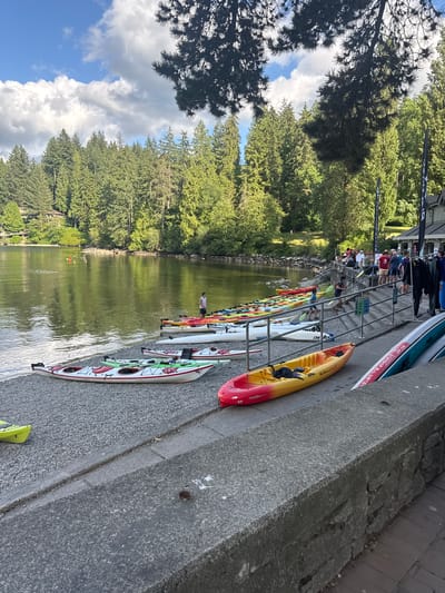 The image depicts an outdoor scene at a waterfront location, likely a recreational facility, on a sunny day with scattered clouds. In the foreground, a concrete barrier runs horizontally across the bottom of the frame. To the left of the barrier, a pebbled shore is visible, lined with numerous kayaks of various colors, including red, yellow, white, and green. Some of these kayaks are positioned in rows, while others are scattered. A bright yellow kayak with "Malibu 11.5" written on it is prominent in the mid-ground, positioned on a paved area.
Beyond the kayaks, a body of water, likely a lake or inlet, reflects the surrounding trees and sky. Several people are present, some standing near a set of stairs leading up to a building. The building appears to be a boathouse or similar facility, with dark siding and a flag attached to its side. A group of people, dressed casually, are gathered on and around the stairs. Further in the background, a dense forest of evergreen trees covers the hills. In the water, a few individuals can be seen in kayaks. The scene suggests a place for water sports and recreation.
Based on the visual cues, the location is likely Deep Cove Park in the District of North Vancouver, British Columbia, Canada. Deep Cove is known for its picturesque waterfront, surrounded by forests and popular for kayaking and other outdoor activities. The presence of numerous kayaks, a boathouse-like structure, and the characteristic evergreen-covered hills strongly suggest this specific area.