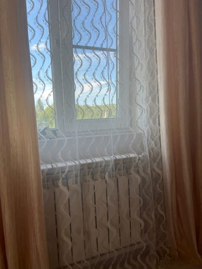 The image shows a window with curtains and a radiator. The window is partly covered by a sheer white curtain with a wavy pattern, and to the left and right are thicker, peach-colored curtains. Through the sheer curtain, a view of trees and a blue sky with some clouds is visible. Below the window is a white radiator with a ribbed surface. The scene is indoors, likely in a room. There are no people or animals visible. The image does not provide any information or context to suggest a specific location in the city of Zhukov, Russia.