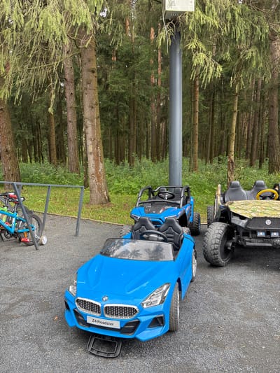 The image depicts a children's play area in a wooded environment, likely a park or recreational facility. In the foreground, a bright blue toy car, identified as a "Z4 Roadster" by its license plate, is parked on a gravel surface. To the right of the blue car, another toy vehicle, a camouflage-patterned all-terrain vehicle (ATV), is visible. Behind these, a smaller blue ATV is also present. To the left of the blue Roadster, a blue bicycle with training wheels is leaning against a metal rack. The background is dominated by a dense forest of tall, slender trees, indicative of a natural, possibly rural setting. The overall scene suggests an outdoor recreational space designed for children's activities. The location is identified as Pavlovskaya Sloboda, Russia.