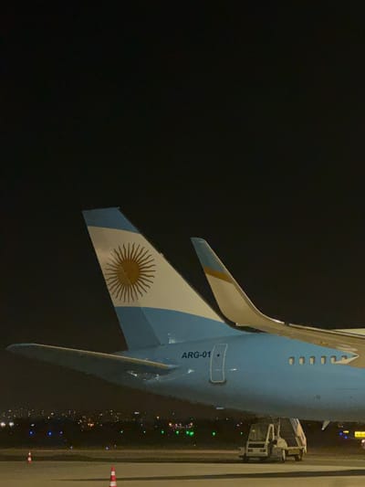 An upward, medium shot depicts the tail fin of the ARG-01 aircraft against a dark, nighttime background. The tail fin is painted with the Argentinian flag, featuring a white field and blue stripes along the top and bottom edges. A golden sun emblem is displayed in the center of the white field. The plane’s body has ARG-01 printed on it, is blue and runs along the right of the frame, extending beyond the top edge. A ramp connects to the plane. Runway edge lights and city lights stretch along the bottom right of the frame, indicating an airport environment at night. The location appears to be at night in Yagel, Israel.