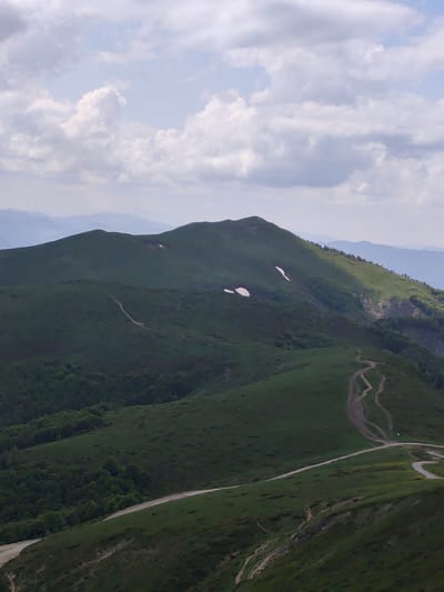 The image displays a vast mountainous landscape in the city of Unknown, Montenegro. The scene is dominated by rolling hills and mountains covered in lush green grass and scattered patches of darker green trees. Several unpaved dirt tracks are prominently visible, winding across the grassy slopes, particularly in the midground and lower right of the frame. Two distinct patches of snow are present on the upper sections of the closer, greener mountains. In the far background, successive layers of mountains appear in desaturated blue and grey tones, indicating atmospheric distance. The sky, occupying the upper portion, is light blue with scattered white and grey cumulus clouds. No human figures, animals, or man-made structures (other than the tracks) are depicted. The overall lighting is natural daylight, diffused by the cloud cover, resulting in soft shadows.