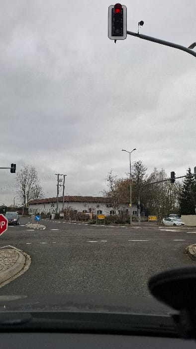 The scene depicts a street intersection in Weiler-la-Tour, Luxembourg, observed from the interior of a stationary vehicle on an overcast day. A traffic light positioned overhead and to the right of the viewer displays a red signal. To the left, a partially visible red octagonal STOP sign stands near the roadside. The asphalt road surface shows painted white dashed and solid lines. In the center of the intersection, a small, raised circular island contains a blue directional sign.
A dark-colored passenger vehicle is visible approaching the intersection from the left. On the right, a white sedan is parked along the roadside. A prominent, two-story building with a light-colored facade and a reddish-brown tiled roof dominates the background, situated behind the intersection. Several bare deciduous trees are interspersed throughout the area, with some evergreen trees on the far right. Multiple streetlights and utility poles with overhead lines are present. No individuals are visible.
