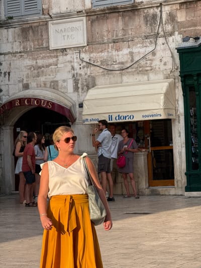 This is a medium shot of a street scene in Split, Croatia, likely in the historic center. In the foreground, a woman with blonde hair and sunglasses is looking to the right. She is wearing a white sleeveless top with ruffled shoulders and a flowing, mustard-yellow skirt tied at the waist. She is carrying a light grey tote bag over her left shoulder. Her left arm is bent, and her hand rests near her waist, showing a bracelet and a ring.
In the mid-ground, several people are standing in front of shops. To the left, there is an arched entrance with a sign reading "MY PRECIOUS." To the right, a beige awning covers a shop with the words "il dono" and "souvenirs" visible. People are seen browsing and interacting within the vicinity of these shops. A man in a striped shirt is standing to the right, and a woman in a patterned dress with a bright pink purse is also visible.
In the background, a stone building with a sign reading "NARODNI TRG" is mounted on the wall. The architecture suggests an older European city. The ground is paved with cobblestones or large flagstones. The lighting indicates it is a sunny day, with distinct shadows cast on the ground.