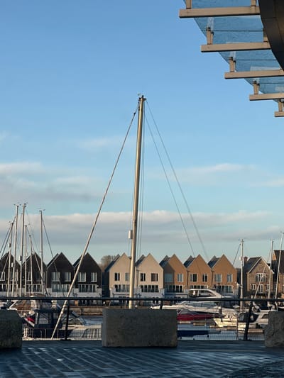 The image depicts a marina scene at Chatham Maritime, Gillingham, United Kingdom, under a clear, light blue sky with scattered horizontal white clouds. In the foreground, a dark, wet-appearing paved surface is visible, extending from the bottom of the frame. Two rough-textured, light grey concrete blocks stand on this surface, flanking a dark metal fence or railing that runs horizontally.
A prominent, light-colored mast of a sailboat stands centrally, extending vertically through the middle of the frame, with multiple thin lines of rigging. To the left and right of this central mast, numerous other masts of various moored sailboats are visible, along with parts of powerboats and sailing vessels docked in calm water behind the railing.
In the mid-ground, a row of modern, multi-story townhouses with distinct gabled roofs lines the waterfront. These houses display varied facades, including dark brown/black, light beige, and medium tan, each featuring multiple windows and some with lower balconies or terraces. A distant line of trees is visible behind the houses on the far right.
In the upper right quadrant, a portion of a contemporary glass and metal canopy structure is present, consisting of horizontal metal beams supporting translucent panels. A small, dark, elongated object, consistent with an aircraft, is faintly visible in the upper-left sky. No people are present in the scene.