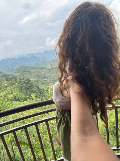 A woman stands on a balcony overlooking a lush, green valley in what appears to be the Quản Bạ Commune area of Vietnam. Her dark, curly hair is prominent in the foreground. She is wearing a light-colored top and green shorts. A dark metal railing lines the edge of the balcony, providing a view of the terraced hills and mountains in the background. The sky is overcast, creating a diffused light across the scene.