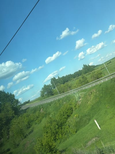 The image depicts a landscape scene, likely a roadside, with a bright blue sky filled with scattered white clouds. A dark, thin line, possibly a wire or cable, diagonally bisects the upper left portion of the sky. Below the sky, a verdant green hillside slopes downwards. A highway with a white guardrail runs along the middle ground, curving to the right. Dense green trees and vegetation cover the lower part of the slope and the area beyond the highway. A single white post with a red cap is visible on the grassy slope in the lower right quadrant. The overall impression is one of a rural or semi-rural environment on a clear day. There are no people or specific identifiable structures in the image that would pinpoint a precise location within the city of Камаево, Russia.