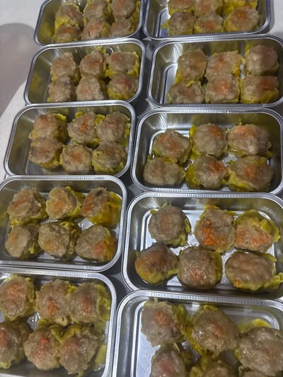 The image shows multiple aluminum trays filled with dim sum. The dim sum are small, steamed dumplings with a meat filling, wrapped in a yellow dumpling wrapper. Some of the dim sum have a reddish-orange topping, possibly from shrimp or carrot. The trays are arranged in rows, suggesting they are prepared for a meal or sale. The lighting suggests an indoor environment, likely a kitchen or food preparation area. The specific location in the city of Kalijaten, Indonesia, cannot be determined from the image alone.