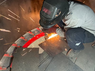 A worker wearing a welding mask and gloves is grinding metal, creating sparks. They are kneeling near a curved metal structure with red markings, surrounded by metal sheets on a concrete floor. The location in the city of Zaljevo, Montenegro is not possible to confirm from the image alone.