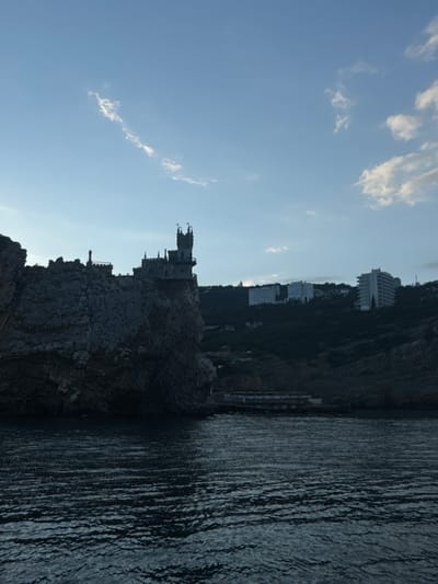 The image shows Swallow's Nest, a decorative castle located in Гаспринский поселковый совет, Ukraine. The castle is situated on a rocky cliff overlooking the sea. Behind the castle, further up the hillside, are several large buildings. The sky is blue with some clouds. The sea is dark and appears to be calm. The image is oriented with the castle and sea on the right side.