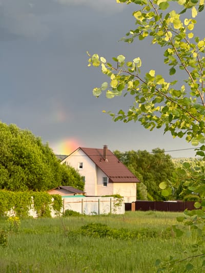 The image shows a rural scene in Vysokinichi, Russia. A small house with a brown roof and white siding is visible amidst lush greenery. A faint rainbow arcs from the dark, stormy sky towards the house. Branches with green leaves hang in the upper left corner, and a fence with vines runs along the right side of the frame. Tall grass is present in the foreground on the right, and trees fill the background. An electrical wire is partially visible near the top center. The photo is oriented with the house appearing sideways.