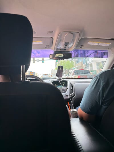 The image shows the view from the back seat of a car, looking forward through the windshield. The sun visors are down, and a smartphone is mounted on the dashboard. A person is visible in the passenger seat, wearing a blue shirt. Another person is in the driver's seat. Outside the car, the scene is a city street with buildings and traffic. The traffic lights are visible, and the buildings have awnings and storefronts. Based on the buildings in the background, it is in Atlanta, United States of America.