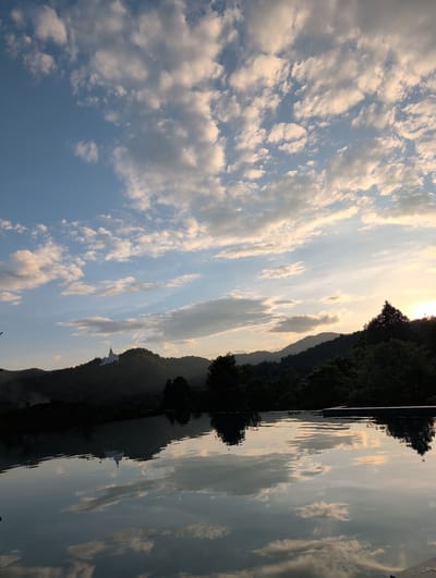 The image depicts a serene waterscape at sunset, with the sky filled with scattered clouds reflecting on the calm water. The scene is set against a backdrop of lush, tree-covered hills, silhouetted by the setting sun. The sun's rays cast a warm, golden glow across the sky and water, creating a peaceful and idyllic atmosphere. In the distance, a white stupa-like structure is visible on the hillside, suggesting a religious or cultural site. The overall mood is tranquil and contemplative, evoking a sense of natural beauty and spiritual calm. The location is identified as Ban Pong Nuea, Thailand. The image appears to be taken from a boat or the water's edge, offering a unique perspective of the surrounding landscape. The time of day is sunset, and the weather is clear with some cloud cover. No text is visible in the image.
