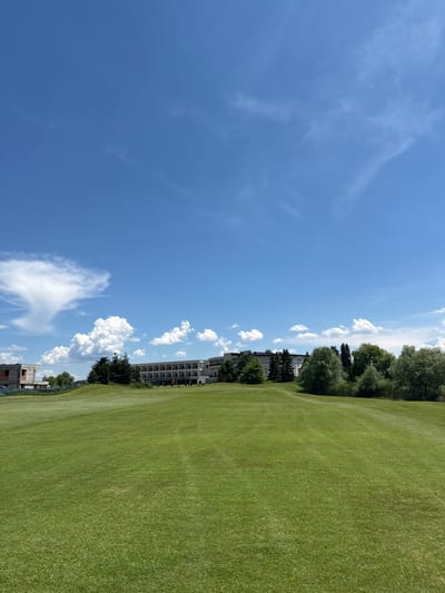 A wide shot captures a meticulously manicured golf course under a bright, blue sky dotted with wispy white clouds. In the middle ground, a long, modern building with a beige facade and numerous windows stands behind a line of lush green trees. To the left of the building, a partially constructed building with exposed brick and scaffolding is visible. The foreground is dominated by the undulating, vibrant green grass of the golf course, with faint tracks suggesting recent mowing. A small, yellow flag, typical of a golf course, is placed in the grass near the building. The scene appears tranquil and expansive, characteristic of an outdoor recreational area. The location is identified as Ravno Pole, Bulgaria.