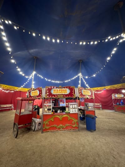The interior of a circus tent, located in Tejalpa, Mexico, is dominated by a large blue canvas dome adorned with strings of lights. Underneath the tent, several vendor carts are arranged, selling items like nachos and tickets. A blue trash can is near one of the vendors. There are no people visible. The ground is a neutral tan-gray.
