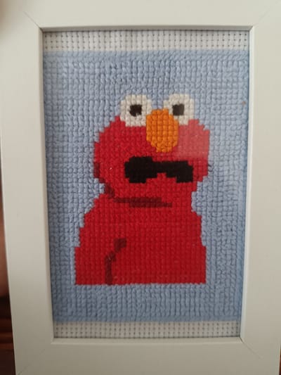 The image shows a framed cross-stitch portrait of the character Elmo. Elmo is depicted in his typical red color, with a black mouth and white eyes with black pupils. The background is a light blue, and the entire piece is framed in a white border. The cross-stitch is on a white Aida fabric, which is visible around the edges of the blue background. The image does not provide any information about the location in Vitoria-Gasteiz, Spain.