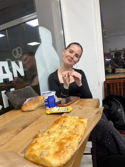 A woman sits at a table indoors, possibly in Strumica, North Macedonia, as the reflections in the glass suggest the Cyrillic letters of the city's name. She is breaking bread with her fingers. On the table, there is a larger flatbread, a small loaf in a bag, a container of yogurt, and a packet. The seating and objects in the background suggest a casual dining establishment.