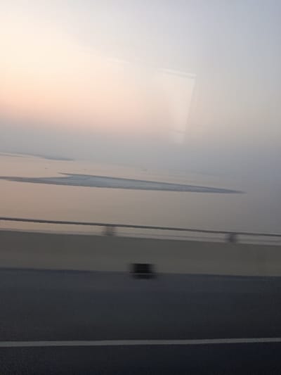 The image depicts an expansive view over the Padma River from the Padma Bridge, located near Naodoba, Bangladesh, likely during dawn or dusk. The scene is characterized by soft, diffused lighting and a hazy atmosphere.

In the foreground, the lower third of the image shows a dark asphalt roadway with a faint, blurred white lane divider line. Above the roadway, a light-colored concrete barrier or parapet wall is visible, topped by a horizontal, dark metal handrail that extends across the frame. A heavily motion-blurred, dark, rectangular object is present on the roadway surface near the base of the barrier.

The midground features a vast body of water, the Padma River, appearing calm with a pale, reflective surface. Within the river, several elongated, lighter-toned landmasses or sandbars are evident, suggesting shallow areas or islands typical of large river deltas. Faint, indistinct outlines of what appear to be distant vessels or small boats are minimally discernible on the water's surface.

The background consists of a hazy sky, transitioning subtly from a warm, pale peach or pink hue near the horizon line to a lighter grey or almost white tone towards the top of the frame. Reflections, possibly from an overhead structure or interior of a vehicle, are faintly visible in the upper portion of the sky, indicating the photograph was taken through a window. No specific individuals or discernible interactions are visible.