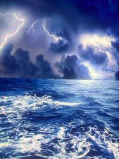 The scene depicts a dramatic and turbulent seascape under a severe thunderstorm, located off the coast of Zelenyi Hai, Ukraine. The upper half of the image is dominated by a dark, heavy sky filled with voluminous storm clouds in shades of deep indigo, grey, and purplish-blue. Multiple bright, jagged lightning strikes illuminate these formations, casting brilliant white and magenta-tinged light across the cloudscape, notably on the left and right sides of the frame. The horizon line is visible as a faint, indistinct demarcation separating the stormy sky from the ocean. The lower half of the image features a rough, agitated sea, with dark blue water churned into numerous whitecaps and foamy crests, particularly prominent in the foreground. The water's surface reflects the intense atmospheric light, highlighting the dynamic movement of the waves. No human subjects or man-made objects are visible within the frame. The environment is one of an open, stormy ocean.