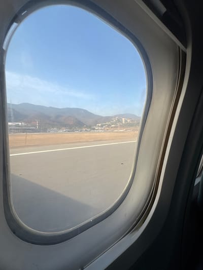 The image shows an aerial view through an airplane window. The runway is visible, stretching towards a town at the base of a mountain range. The mountain range appears to be located in Catia La Mar, Venezuela, judging by the local architecture. There are no people visible in the window's view.