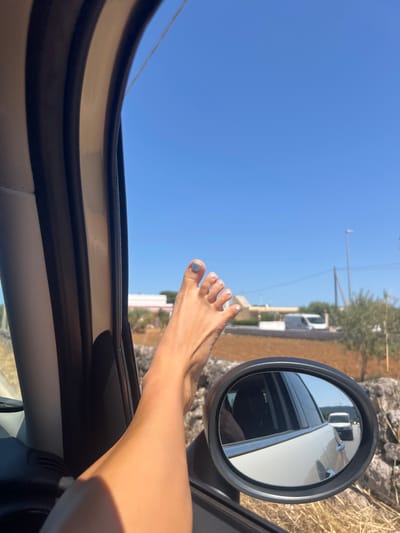 A close-up, eye-level shot shows a person's bare foot with light blue painted toenails resting on the exterior of an open car window. The foot extends out of the frame from the bottom left. The car's interior, including dark and light trim and a side mirror, is visible. The side mirror reflects the road behind, showing a white van and another vehicle. The background is a clear blue sky with a hint of a dry, rural landscape featuring a white building, a street lamp, olive trees, and a stone wall. The scene is likely in Fasano, Italy, given the rural, Mediterranean context.