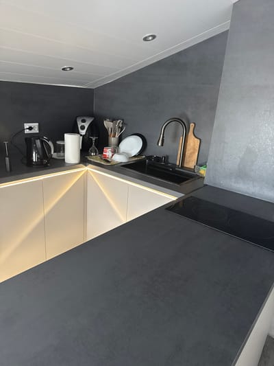 The image shows a modern kitchen counter with a dark gray countertop. On the counter, there is a black electric kettle, a roll of paper towels, a wine glass, and a small appliance that appears to be an air fryer. To the right, there is a black sink with a silver faucet and a wooden cutting board. A stovetop with two burners is visible on the right side of the counter. The kitchen has dark gray walls and a white ceiling with recessed lighting. The cabinets below the counter are white and feature LED strip lighting that casts a warm yellow glow. The scene is devoid of any people or active events. The image is likely a photograph of a kitchen in a residential setting.