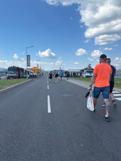 The image depicts an outdoor scene on a sunny day with scattered clouds. A group of people are walking on an asphalt road marked with white lines, suggesting it might be a pedestrian walkway or a closed-off road for an event. On the left side of the road, several food trucks are parked, displaying various signs and banners. One food truck is red and black, with "FOOD TRUCK" clearly visible on its side. Further down the road, there are tents and more food vendors. On the right side of the road, two men are walking away from the camera, one wearing an orange t-shirt and denim shorts, and the other in a dark tank top and shorts. The man in the orange shirt has a large tattoo on his leg. The environment appears to be an open area, possibly a parking lot or event ground, with a fence visible in the background. The sky is blue with cumulus clouds. The location is specified as Dospey, Bulgaria.