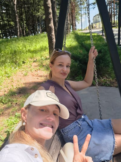 Two women are seated on a swing in a park-like setting. The woman in the foreground is wearing a white cap and a white shirt. She is smiling at the camera and making a peace sign with her right hand. The woman in the background is wearing a purple top and denim shorts. She is also looking at the camera and appears to be smiling. The swing is made of metal chains and is suspended from a dark metal frame. The background shows a wooded area with tall trees, green grass, and a paved pathway. In the distance, a modern building with large windows is visible. The location is identified as Arseniev, Russia.