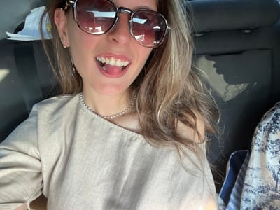 A young woman with blonde hair and sunglasses is taking a selfie in the passenger seat of a car. She is smiling with her tongue sticking out slightly. She is wearing a light-colored shirt and a pearl necklace. The background is the interior of the car, with the headrest visible behind her. The image appears to have been taken in Gelemenovo, Bulgaria.