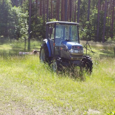 A blue and silver tractor with a cab sits in a grassy field with trees in the background. The tractor is facing right and appears to be stationary. A rotary mower is attached to the rear of the tractor. A fence is visible behind the tractor. The scene is set in Garkalne, Latvia.