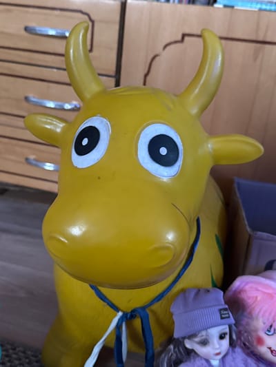 The image features a close-up of a yellow, inflatable, toy horse head, likely a bouncy ride-on toy. The head is prominently displayed, with cartoonish eyes that have black pupils surrounded by white. The horse has short, rounded horns. A blue rope is tied around the horse's neck. Partially visible behind the toy horse are several dolls and a cardboard box. The background includes a light brown cabinet or wall with vertical grooves. The location, Дойная, Russia, is not visually discernible from the image.