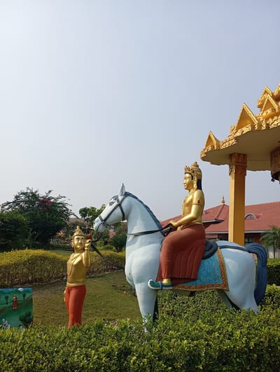 The image shows two statues in a garden in Lumbini Sanskritik, Nepal. One statue depicts a person riding a white horse, while the other statue stands next to the horse's head, holding the reins. Both statues are adorned with gold-colored headwear and reddish-brown clothing. The background includes green bushes, trees, a golden-colored structure, and a red-roofed building. The sky is clear and pale blue.