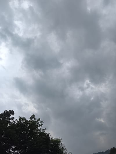 The image shows a dark, overcast sky filled with heavy, grey clouds. The bottom portion of the image is dominated by the dark silhouette of lush green trees. A distant, faint outline of hills or mountains can be seen on the far right, partially obscured by the trees and the low light conditions. The scene appears to be set in an outdoor environment, likely in a natural or semi-natural area within Gunung Malang, Indonesia, given the presence of dense foliage and hilly terrain. No people or specific actions are depicted, the focus is on the atmospheric conditions and the natural landscape.