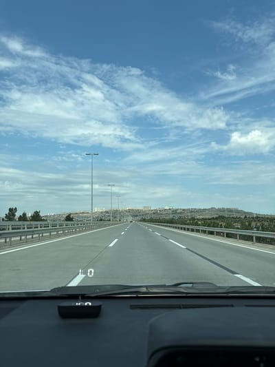 The image shows a road scene in Surakhany Raion, Azerbaijan, taken from inside a vehicle. The perspective is from the driver's seat, looking forward along a multi-lane highway. The road is paved and has clear white lane markings. On the right side of the road, a metal guardrail runs along the edge, separating the highway from the surrounding landscape. Beyond the guardrail, there is a hillside covered in vegetation and buildings, suggesting an urban or suburban environment. In the distance, more buildings are visible, indicating a densely populated area.

The sky is blue with scattered white clouds, suggesting fair weather. The lighting indicates it is daytime, likely late morning or early afternoon given the brightness and the clear sky. Several tall streetlights line the side of the road.

On the dashboard of the vehicle, a black electronic device is visible. There is also a road marking on the asphalt that appears to say "LO" and then "0." The overall mood is one of travel and observation.
