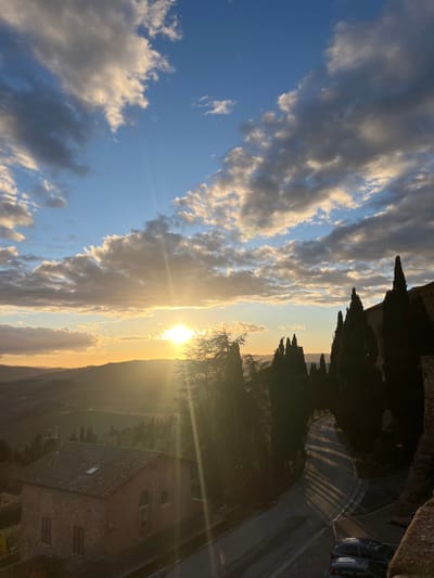 The image presents a serene and picturesque sunset or late afternoon scene, characteristic of the Tuscan countryside near Pienza, Italy. The sky is a blend of clear blue and dramatic clouds; some appear fluffy and bright white-gold, warmly lit by the sun, while others are darker, dense grey formations.

The sun, low on the horizon, is a brilliant, almost blinding golden orb, casting strong rays and a visible sunburst effect across the landscape. Its intense light bathes the entire scene in a warm, golden glow. In the mid-ground, a series of gently rolling hills or mountains are partially silhouetted against the bright sky, extending across the frame.

Tall, slender, dark trees, reminiscent of Italian cypresses, stand prominently along a ridge on the right side of the image, their forms sharply contrasting with the luminous sky. Below these trees and further to the right, a traditional stone or brick building with a distinctive tiled roof comes into view. This structure, likely a house or farmhouse, features multiple windows and what appears to be a skylight on its roof, adding to the charm of the rural setting.

A curved road with painted markings is visible on the far right, leading towards the distance, and a light-colored car is parked near the top right edge. Beyond the building, green fields or cultivated land stretch towards the distant hills. There are no people visible in the image, nor is there any discernible text. The overall impression is one of peaceful natural beauty at golden hour in the Italian countryside.