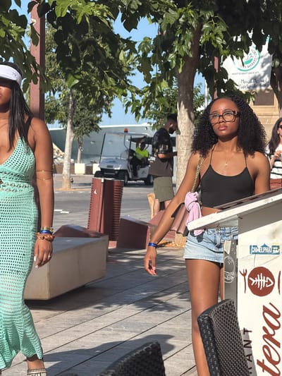 Two women are standing outdoors, in what appears to be a pedestrian area in Eivissa, Spain. The woman on the left is wearing a white visor, a light blue crochet dress, and a gold arm cuff. The woman on the right is wearing glasses, a black tank top, and denim shorts. She is holding a pink purse. Behind them, there are trees, a golf cart, and various structures, including a sign that reads "CAMELPHAT" and part of a restaurant menu. The ground is paved with wooden planks. The sun is shining, casting shadows.