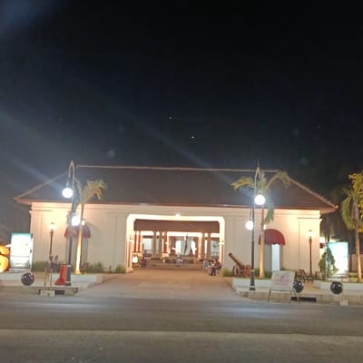 The image captures a nighttime scene at the entrance of a building in Besuki, Indonesia. The building features traditional Indonesian architecture with a wide, open entrance leading into a courtyard. The facade is white with a dark-tiled roof and red awnings over some of the windows. Several palm trees are visible, illuminated by streetlights.

In the foreground, a road is visible, and to the left, a large, illuminated advertising display stands. Two spherical, dark objects on posts are positioned near the building's entrance. To the right of the entrance, a historical cannon is displayed on a raised platform.

Inside the entrance, several people are seated on benches, illuminated by the interior lights. More lights hang from the ceiling of the entrance hall. Further inside, the courtyard appears to be open with more seating and some foliage.

The time of day is clearly night, with a dark sky. The lighting suggests a warm and inviting atmosphere, despite the darkness. The presence of the cannon and the architectural style hint at a historical or cultural site, possibly a museum or a palace complex. The overall impression is one of a well-maintained public space that is open and accessible in the evening. There is no visible text that can be clearly deciphered.