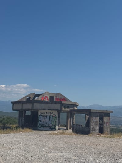 The image shows the remnants of a concrete structure covered in graffiti, located on a hilltop overlooking a valley in Liješnje, Montenegro. The structure appears to be a small, open-air building or shelter with a flat roof and some standing walls. The concrete is weathered and in disrepair, with visible signs of age and neglect.

The graffiti on the structure is varied in style and color, with some tags and larger pieces covering most of the visible surfaces. In the foreground, a gravel path leads up to the structure. The surrounding environment is dry and somewhat barren, with scattered dry grasses and some vegetation on the slopes of the valley. In the background, rolling hills and mountains are visible under a clear blue sky with a few wispy clouds. The overall impression is one of an abandoned and decaying man-made structure in a natural, somewhat remote setting.